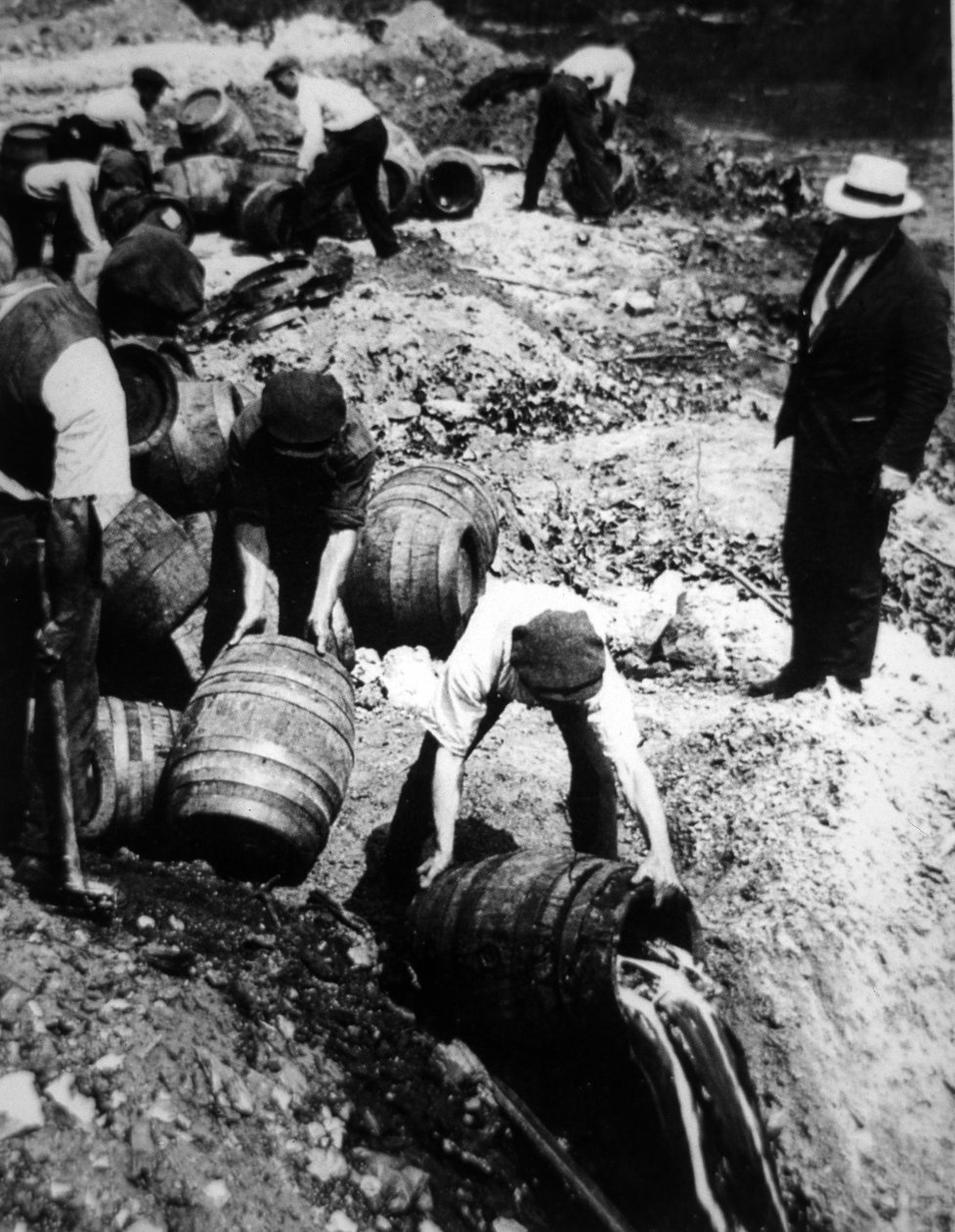 एक अमेरिकी संघीय एजेंट अमेरिकी निषेध युग (1920-33) के दौरान बीयर केगों के विनाश की देखरेख करता है (बी/डब्ल्यू फोटो) द्वारा अमेरिकी फोटोग्राफर