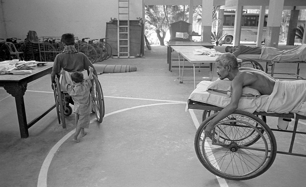 व्हील-चेयर पर एक शारीरिक रूप से विकलांग व्यक्ति को उसके बेटे द्वारा ले जाया जाता है; उसी दुर्दशा के साथ एक अन्य व्यक्ति बांग्लादेश के सावर, ढाका में सीआरपीएल के पुनर्वास के केंद्र में इसे देख रहा है। 2005। द्वारा अज्ञात अज्ञात