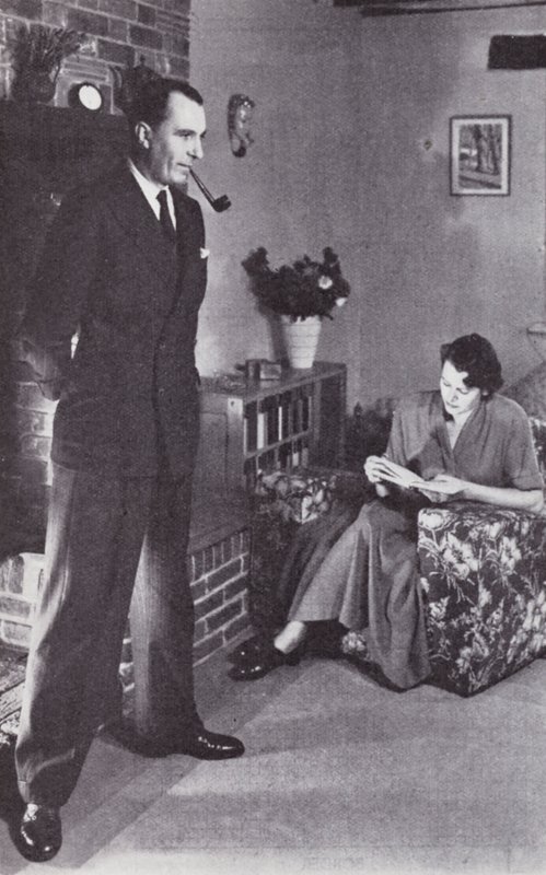 पत्नी पढ़ती है, जबकि पति आग के खिलाफ अपनी पीठ गर्म करता है, c.1950 द्वारा अंग्रेजी फोटोग्राफर